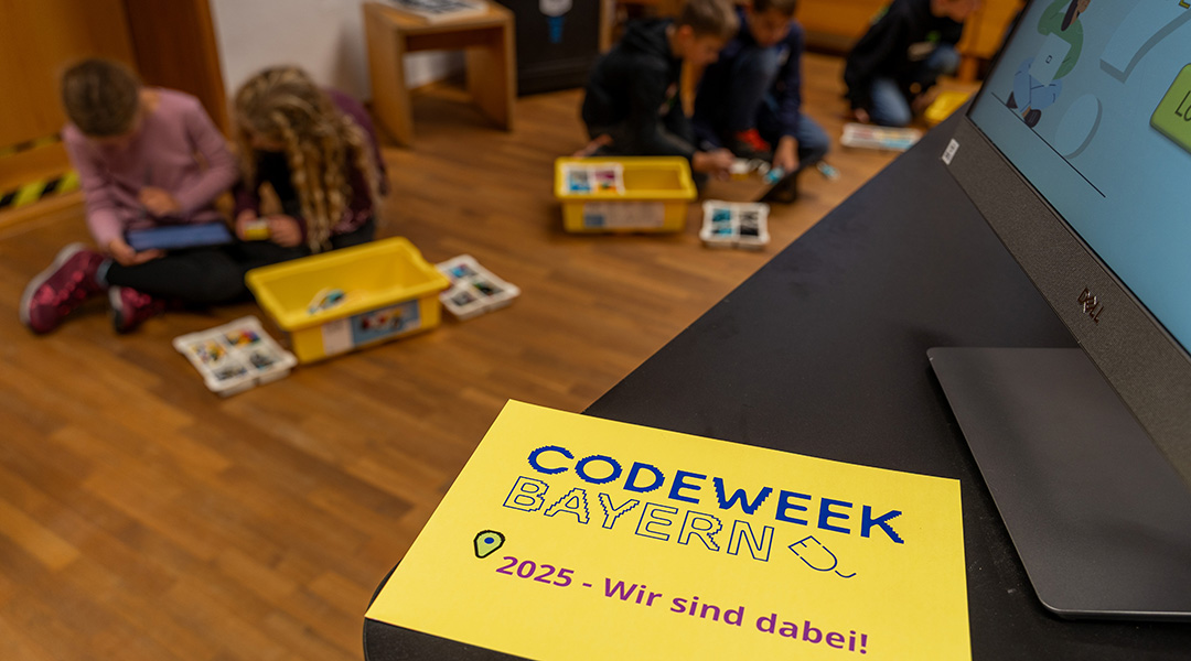 Foto mit Blick in einen Raum, im Hintergrund sind unscharf mehrere Kinder zu sehen, die am Boden sitzen und gerade an LEGO Robotern bauen, im Vordergrund ist ein gelber Zettel zu sehen auf dem 